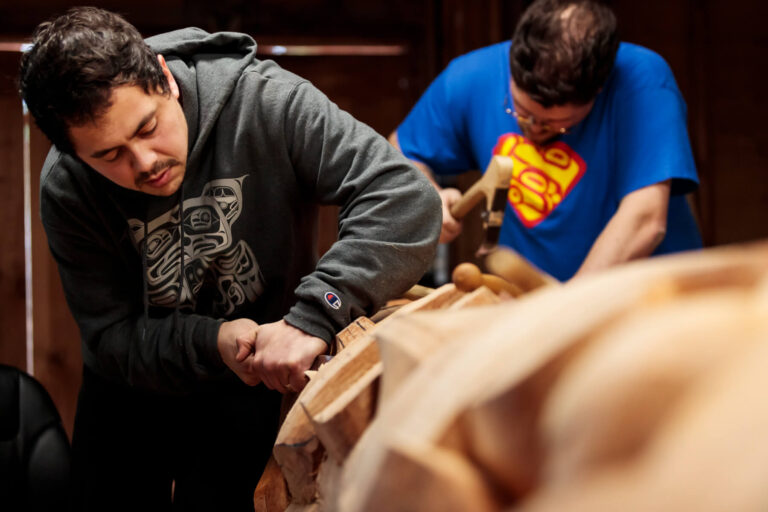 Two men carving a totem pole.